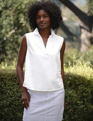 A woman with curly hair stands outdoors, wearing the FRANK & EILEEN Olivia Popover Henley Tank in Famous Denim White paired with light striped pants, looking to the side with a slight smile. The background features blurred green foliage.