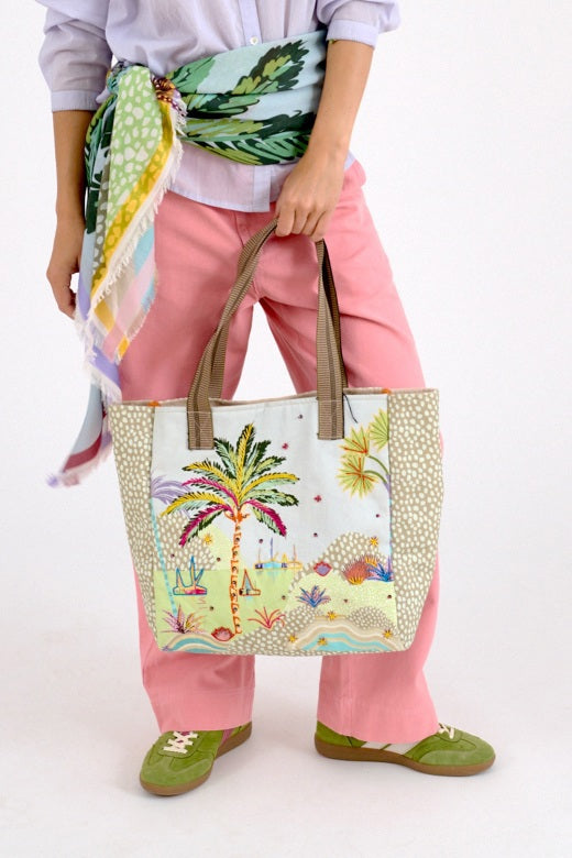A person in pink pants, a colorful scarf, green shoes, and a light shirt holds the STORIATIPIC - CALI TOTE BAG by STORIATIPIC, an embroidered cotton bag with a palm tree and beach scene. The image is cropped above the shoulders.