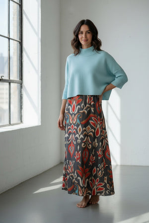A woman with long blonde hair wears the BRITON COURT - SOPHIA Oversized One Size Sweater in Sea Glass by BRITON COURT, featuring three-quarter sleeves, paired with a colorful patterned skirt, standing against a light gray background.