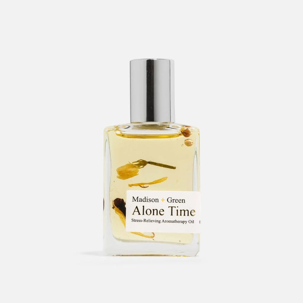 A clear glass bottle with a silver cap holds yellow relaxing body oil infused with small flower petals. The label reads “MADISON AND GREEN - ALONE TIME RELAXING AROMATHERAPY BODY OIL MINI,” highlighting an essential oil blend by MADISON GREEN crafted to help you unwind and relax.