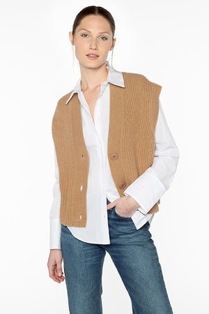 A person stands against a plain background wearing a white button-up shirt with rolled-up sleeves, the KINROSS Rib Button Knit Vest in brown, and blue jeans. The person has a neutral expression, with one hand in their pocket and hair pulled back.