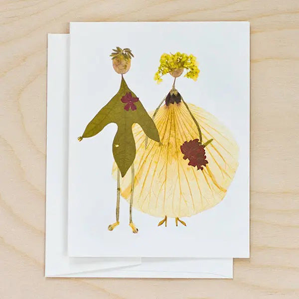 The PETAL PEOPLE PRESS - BETROTHED CARD by PETAL PEOPLE PRESS features two figures in formal wear crafted from real leaves and petals. Shown on a white surface with clippers, greenery, and paperclips nearby.