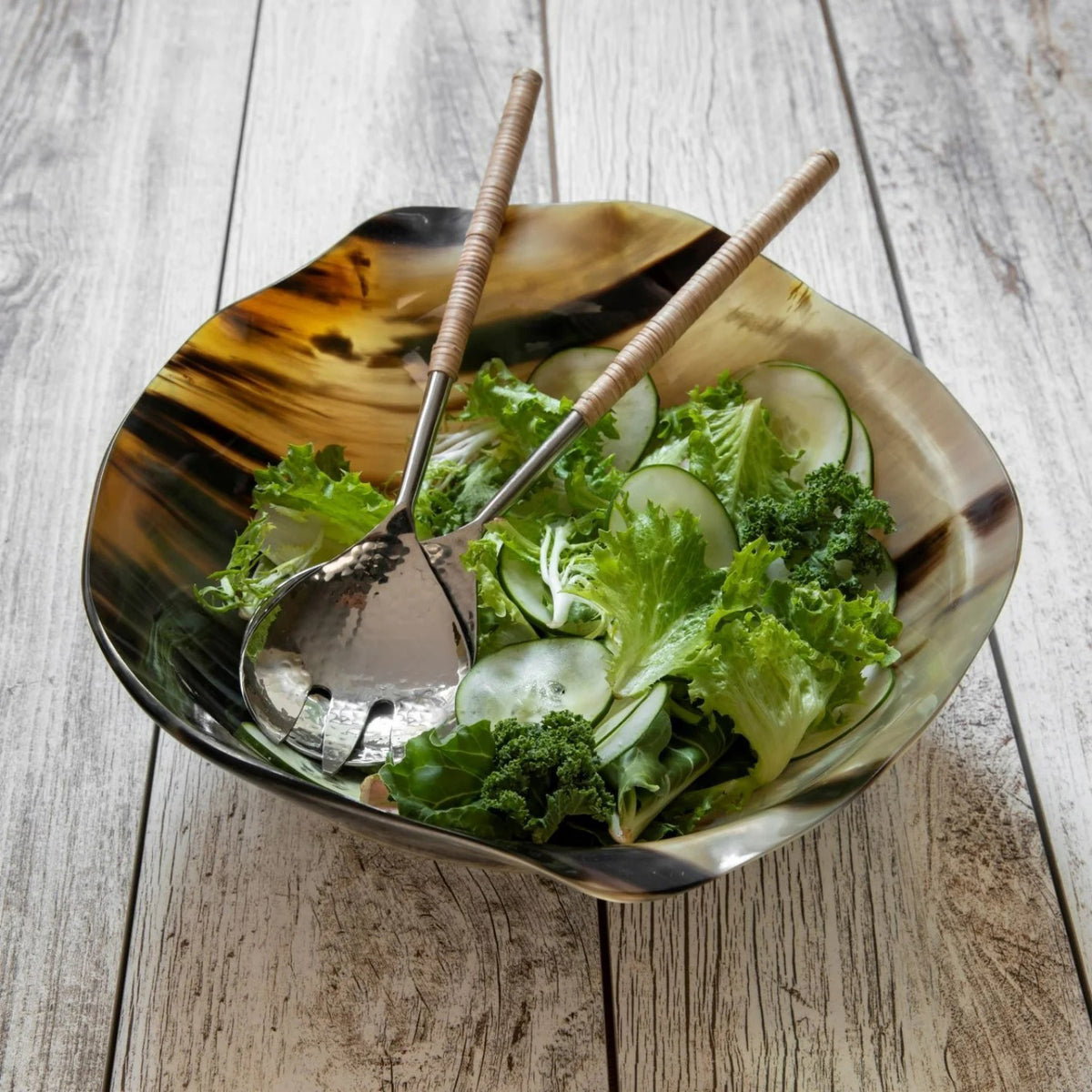 The HORN WAVY ROUND BOWL by BE HOME, INC, ethically crafted from water buffalo horn, showcases a polished, reflective surface. Its irregular, organic shape rests on a flat blue background. The natural blend of black and tan colors highlights the bowl&#39;s unique texture and sheen.