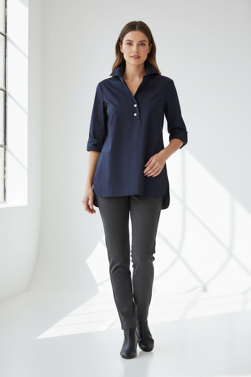 A woman wearing the HINSON WU Aileen Tunic Shirt—a navy blue button-up with three-quarter sleeves and tunic-length—paired with dark gray pants, stands against a white wall, facing slightly to the side.