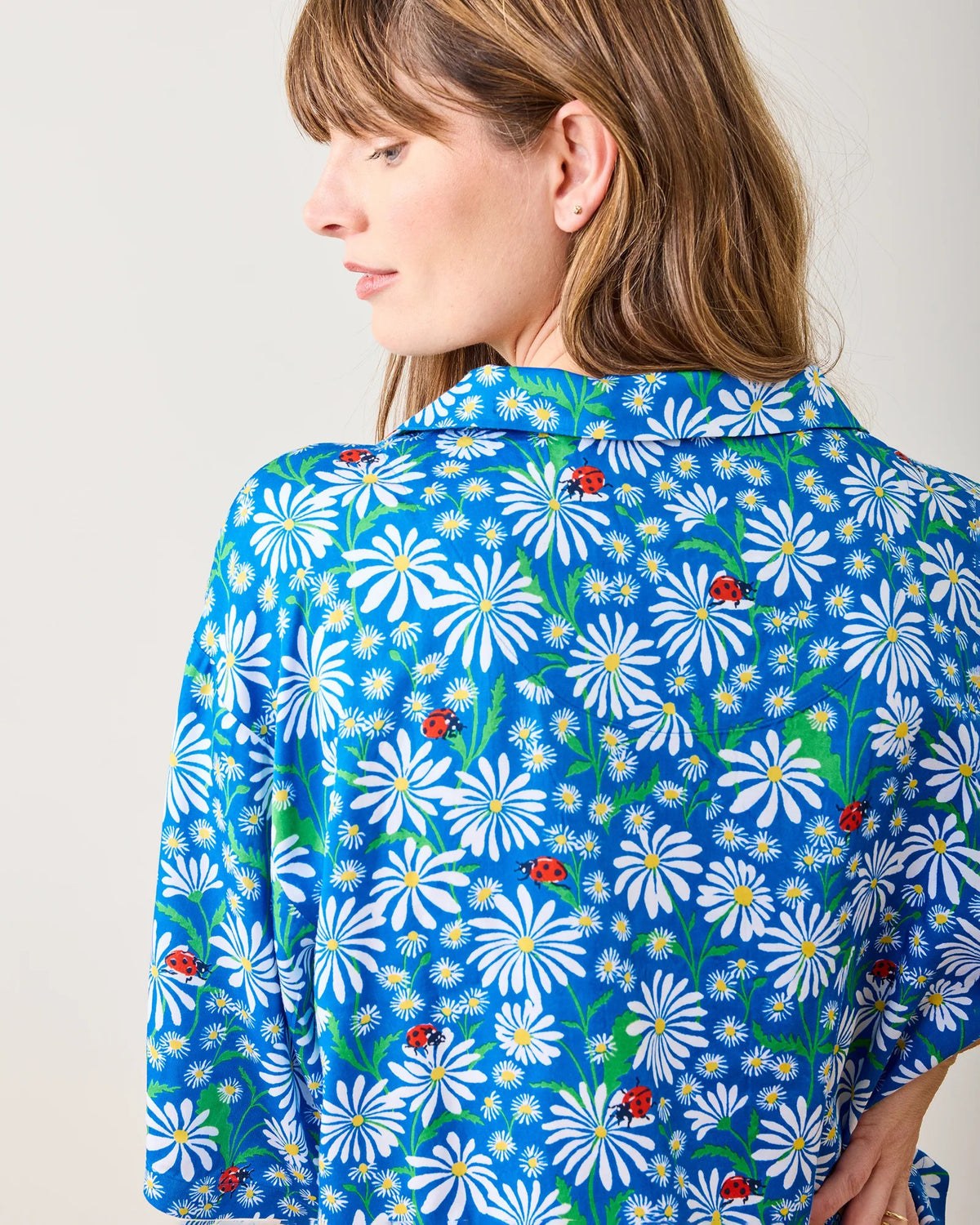 Wearing the PRINTFRESH Satin Lucky Daisy Pajama Short Set with a blue floral design of white daisies, a person with long brown hair and bangs stands barefoot on a wooden floor, leaning against a light-colored wall in a relaxed posture.