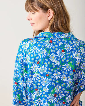 Wearing the PRINTFRESH Satin Lucky Daisy Pajama Short Set with a blue floral design of white daisies, a person with long brown hair and bangs stands barefoot on a wooden floor, leaning against a light-colored wall in a relaxed posture.