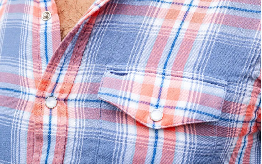 Close-up of a person's chest in the CRIQUET - DOUBLE CLOTH FLANNEL PEARL SNAP SHIRT IN SUNSET, a blue, white, and orange plaid flannel with pearl snaps, front pocket with flap, and visible chest hair at the open collar.
