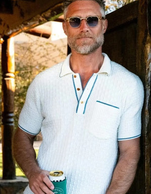 A smiling gray-haired man in sunglasses and khaki pants wears the CRIQUET Bungalow Sweater Polo in White, featuring a blue-trimmed collar and pocket, standing against a plain background.