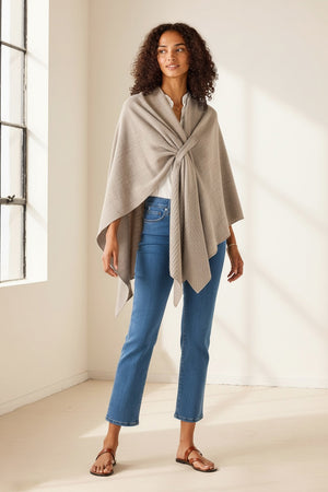 A woman stands indoors in a bright room with light streaming through a large window, smiling softly. She wears her natural curly hair loose and is styled in blue jeans, brown sandals, and a white shirt layered with the MERSEA - MODERN TRAVEL WRAP IN DRIFTWOOD by MERSEA, featuring a securing loop.