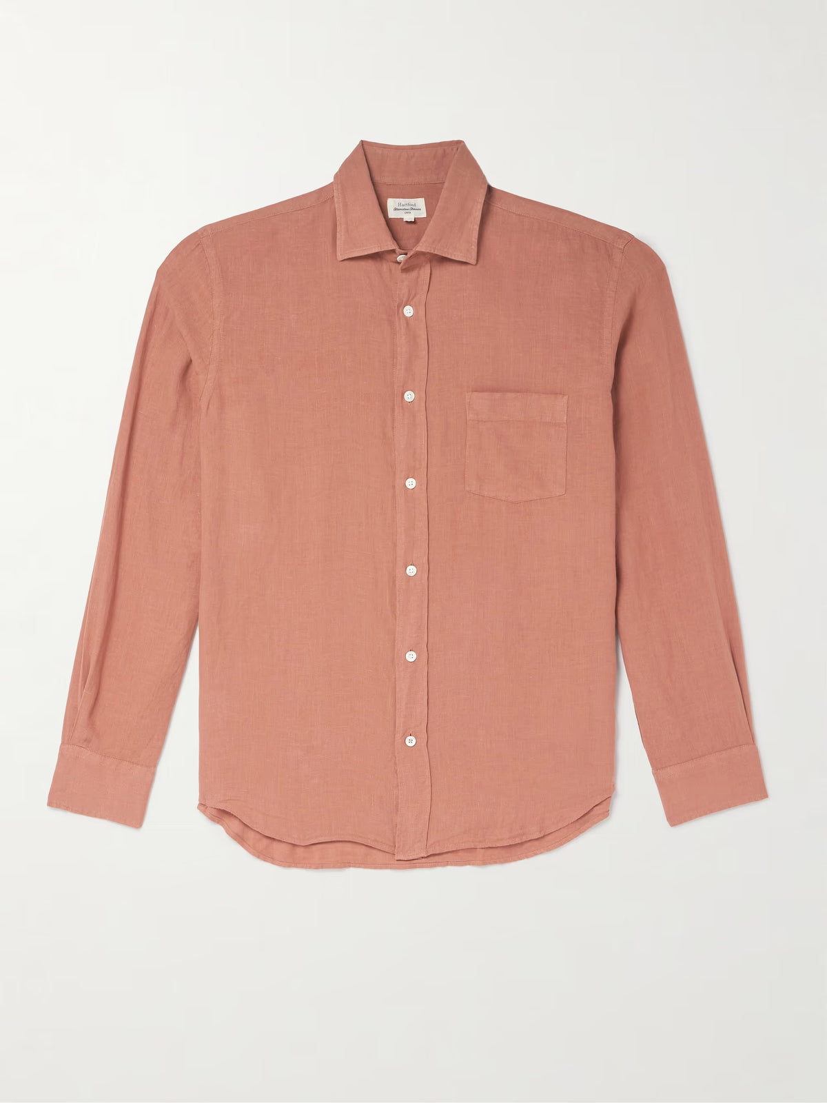 A person wears the HARTFORD - PAUL LINEN SHIRT IN FADED COPPER by HARTFORD, paired with light pants, standing with one hand in their pocket against a plain background.