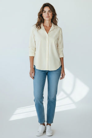 A woman with wavy brown hair smiles slightly, wearing the FRANK & EILEEN EILEEN Relaxed Button Up Shirt in Italian Cotton Thin Yellow Stripe, paired with blue jeans and white sneakers. She stands in a plain, well-lit background.