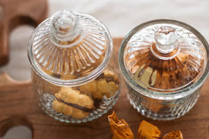 ETUHOME Bistro Glass Biscuit Jars with lids sit on a wooden tray, filled with snacks and orange-wrapped candies. The classic jar design by ETUHOME stands out against the softly blurred background.
