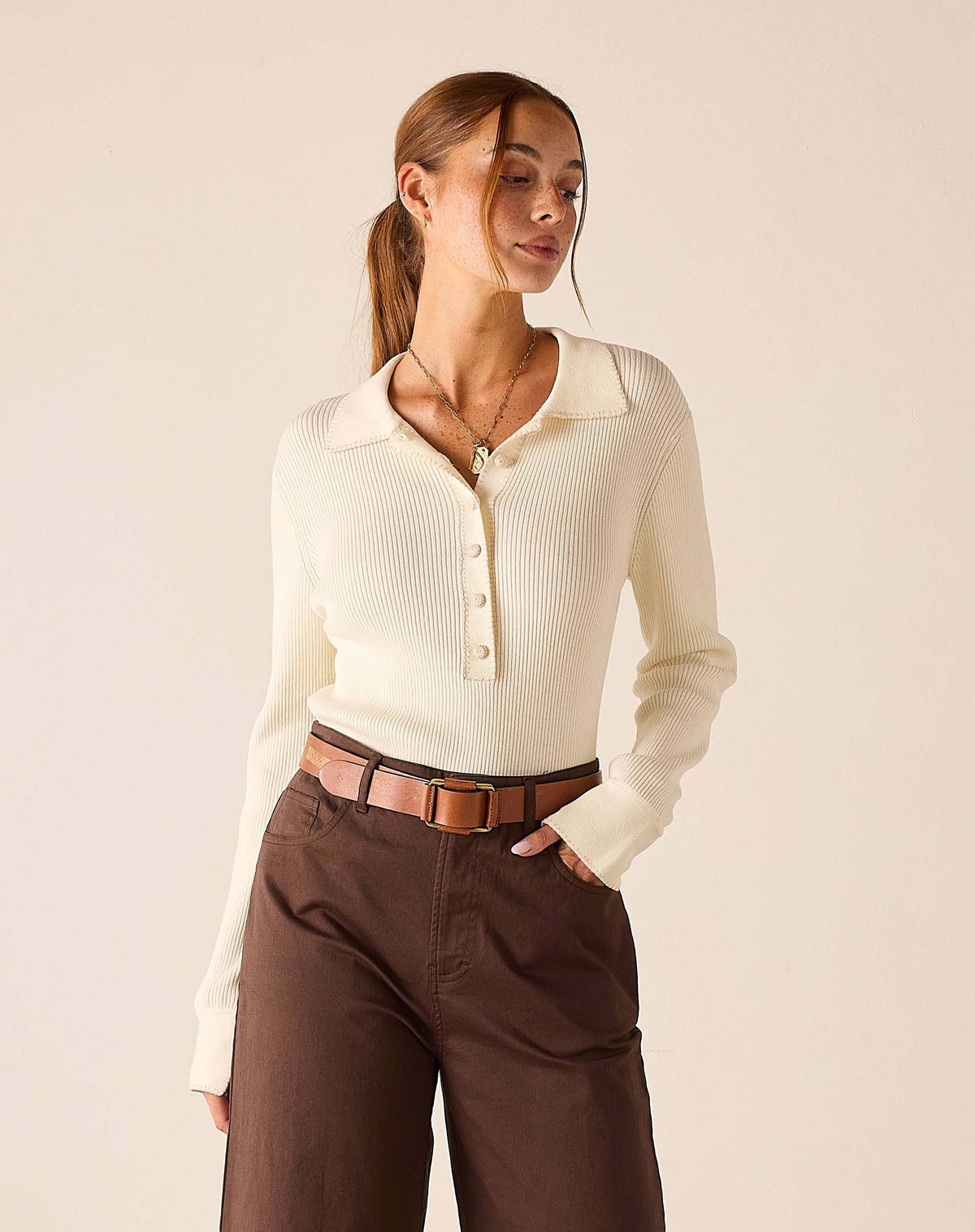 A woman stands barefoot indoors next to a large sunlit window, wearing the MERSEA - LUDLOW LONG SLEEVE POLO IN CREME with a collared neckline, paired with high-waisted brown wide-leg pants and a belt.