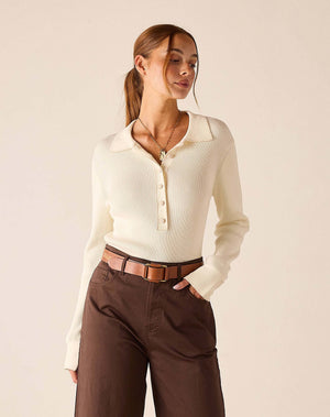 A woman stands barefoot indoors next to a large sunlit window, wearing the MERSEA - LUDLOW LONG SLEEVE POLO IN CREME with a collared neckline, paired with high-waisted brown wide-leg pants and a belt.