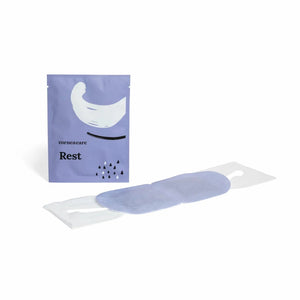 Hands hold a CORNEACARE - REST Self Heating Warm Compress as one hand removes a gray eye patch. The light blue pouch's minimalist design highlights its soothing eye dryness relief, enhanced by black and white elements on a plain backdrop.