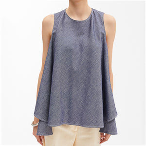 A model wears the MEIMEJ - SLEEVELESS DENIM TUNIC by MEIMEIJ, a tunic-length, light cotton denim piece with side slits, styled with light-colored pants and a silver bracelet, shown in profile against a plain white background.