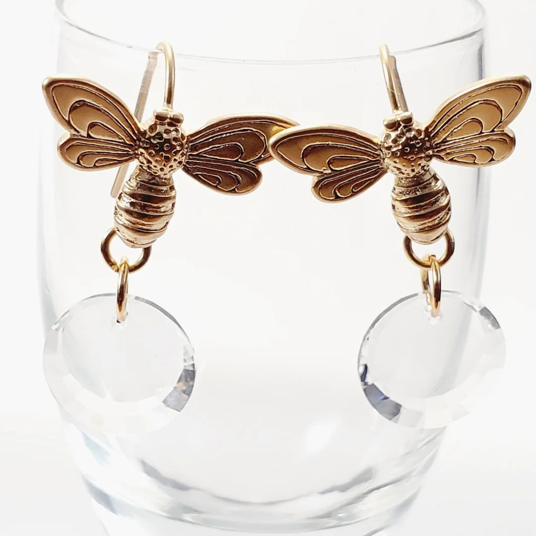 The VERDIER - HONEY BEE FROP EARRINGS WITH SWAROVSKI MOON DISC EARRINGS by VERDIER, featuring gold bee shapes with clear Swarovski crystal drops, are displayed hanging on the rim of a clear glass.