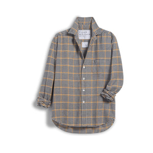 A blonde woman stands indoors, smiling softly, wearing the FRANK & EILEEN EILEEN Relaxed Button Up in Italian Flannel Camel Black Plaid and black pants. The cozy room features a fireplace, wooden floors, a plant, and stacked books on a table.