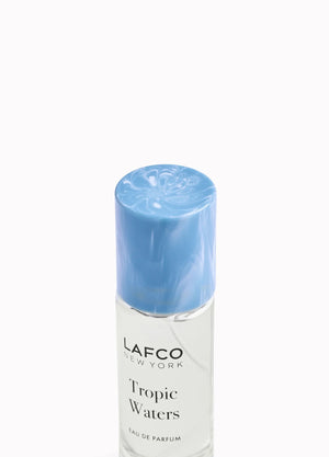 A LAFCO FINE FRAGRANCE, TROPIC WATERS 10ML box stands upright in a halved coconut, water splashing around it against a neutral background—an evocative display for this long-lasting scent inspired by the essence of coconut water.