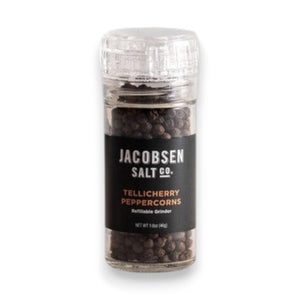 A clear glass grinder filled with black peppercorns rests on a light surface. The label displays “Jacobsen Salt Co.” and “Tellicherry Peppercorns” in white and gold text. Behind, a plain light gray wall forms the background. This is the JACOBSEN SALT - TELLICHERRY PEPPERCORNS WITH GRINDER by JACOBSEN SALT CO.