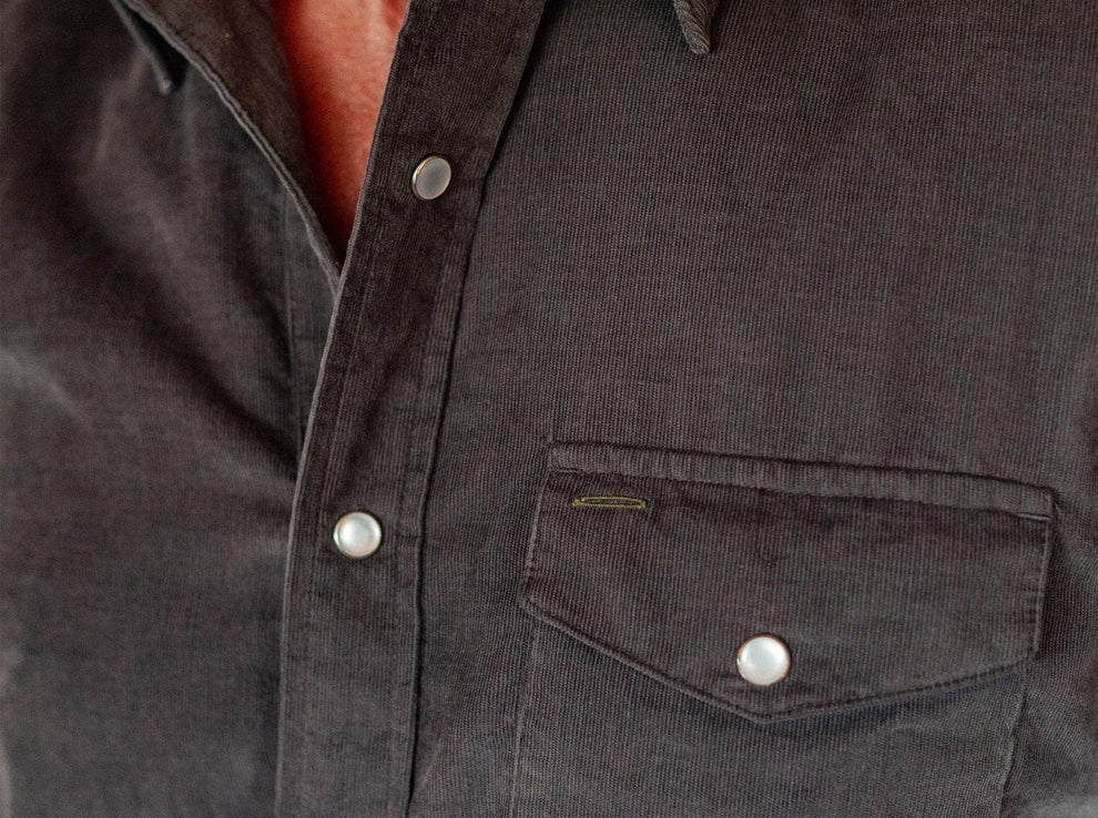 A smiling man in a brown hat and the CRIQUET Short Sleeve Corduroy Pearl Snap Shirt in Iron Horse holds a canned drink in his right hand, standing against a white background.