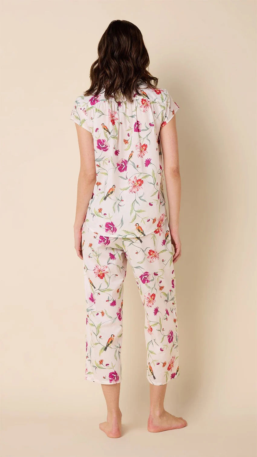 A person smiles while wearing the CAT&#39;S PAJAMAS - ROSA AND BIRD LUXE PIMA CAPRI PAJAMA SET, featuring a floral design on 100% cotton with short sleeves and capri pants. The fabric is light in color with colorful flowers, balancing comfort and style against a plain, light background.