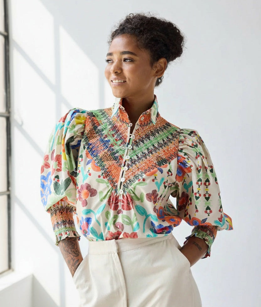 A woman smiles and looks to the side, hands in her cream pants pockets, wearing the HOPE FOR FLOWERS Smocked Shirt in Custard Floral by HOPE FOR FLOWERS against a plain light background.