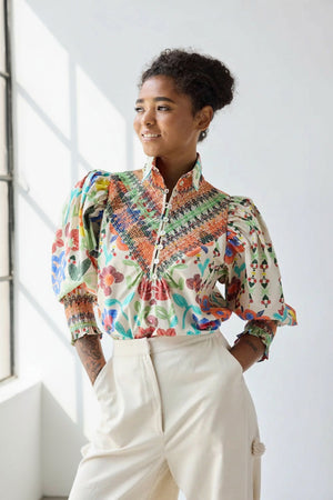 A woman smiles and looks to the side, hands in her cream pants pockets, wearing the HOPE FOR FLOWERS Smocked Shirt in Custard Floral by HOPE FOR FLOWERS against a plain light background.