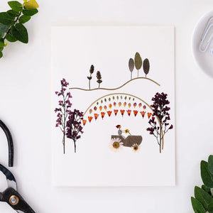 The PETAL PEOPLE PRESS - FARMER IN THE DELL CARD by PETAL PEOPLE PRESS features a car driving under a rainbow of hearts, surrounded by flowers, trees, and hills—an ideal all-occasion card for any nature lover.