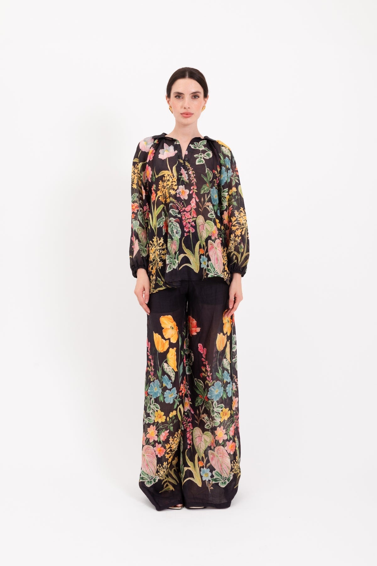 A person stands facing forward, wearing the LAVI - APHRODITE BOTANICAL BLOUSE by LAVI—a loose-fitting, long-sleeved statement blouse featuring a colorful floral pattern—against a plain white background.