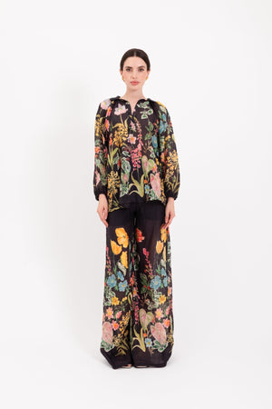 A person stands facing forward, wearing the LAVI - APHRODITE BOTANICAL BLOUSE by LAVI—a loose-fitting, long-sleeved statement blouse featuring a colorful floral pattern—against a plain white background.