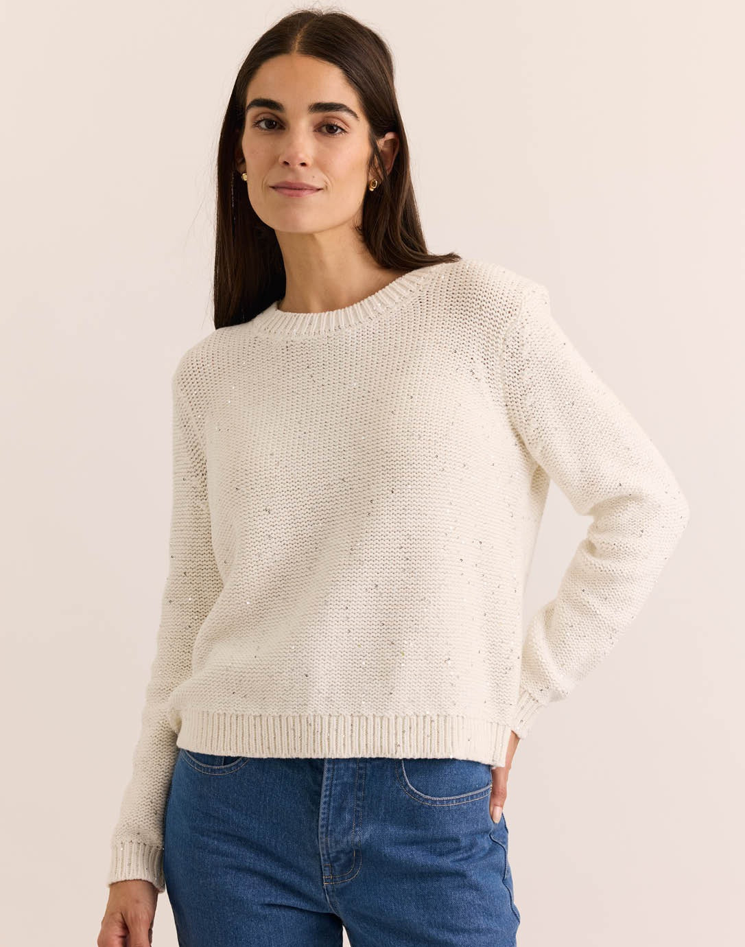 A woman with long brown hair wears the HINSON WU - QUEENIE Long Sleeve Sequin Sweater in Pearl White and blue jeans, standing against a plain light background and looking slightly to the side with a relaxed expression.