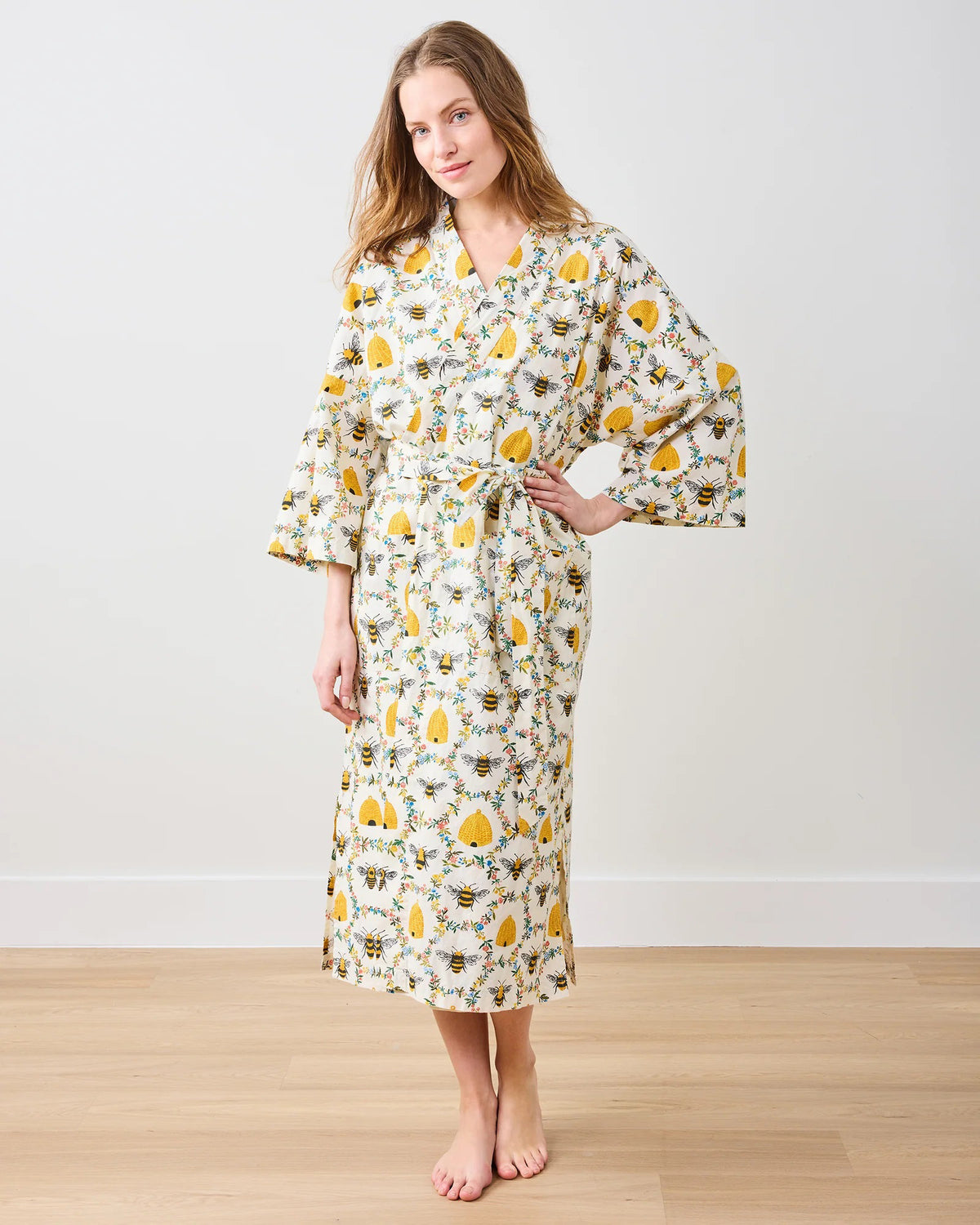 A person models the PRINTFRESH - SWEET BEE COTTON ROBE, featuring a floral design with sunflowers and leaves. Made from organic cotton, it has long sleeves and a waist tie, set against a plain white wall.