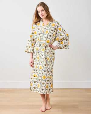 A person models the PRINTFRESH - SWEET BEE COTTON ROBE, featuring a floral design with sunflowers and leaves. Made from organic cotton, it has long sleeves and a waist tie, set against a plain white wall.