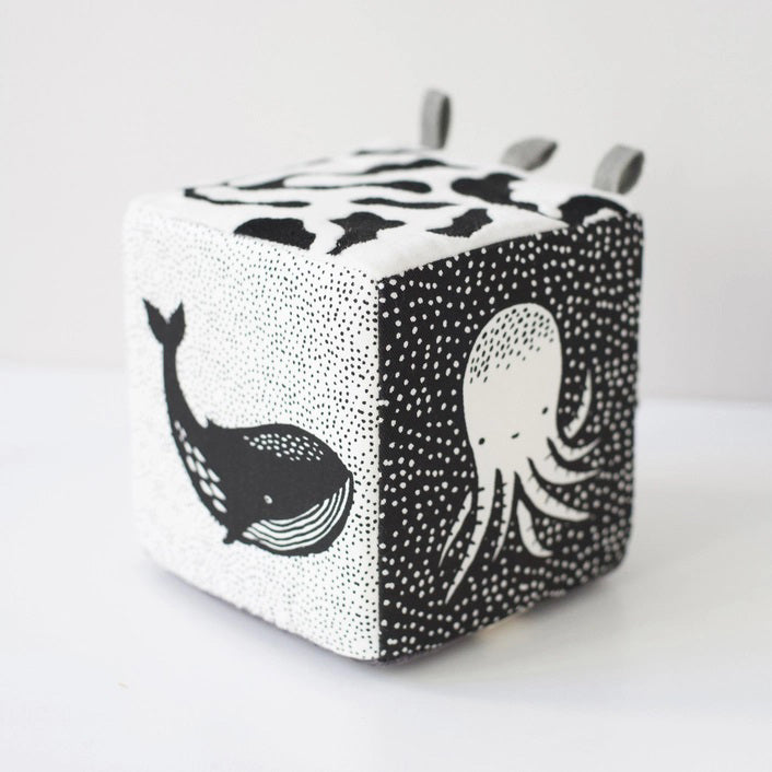 The WEE GALLERY - OCEAN SOFT BLOCK is a monochrome plush cube baby toy featuring high-contrast black and white patterns. Each side of the cube showcases illustrations of ocean life, including a whale and an octopus. Made from soft organic cotton, it also has fabric loops on one corner, making it ideal for infants and toddlers.