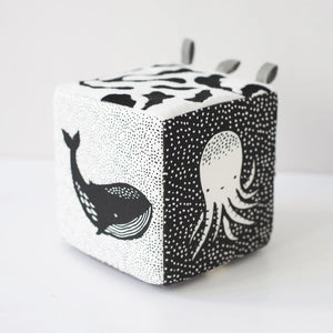 The WEE GALLERY - OCEAN SOFT BLOCK is a monochrome plush cube baby toy featuring high-contrast black and white patterns. Each side of the cube showcases illustrations of ocean life, including a whale and an octopus. Made from soft organic cotton, it also has fabric loops on one corner, making it ideal for infants and toddlers.