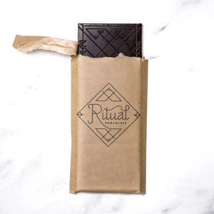 The image shows a light brown rectangular package of Ritual Chocolate labeled "RITUAL CHOCOLATE - BOURBON BARREL AGED CHOCOLATE BAR 75%." This limited edition chocolate, crafted from aged cacao nibs, features geometric designs in silver and white and contains 2.12 oz (60g) of chocolate. The background is a marble surface.