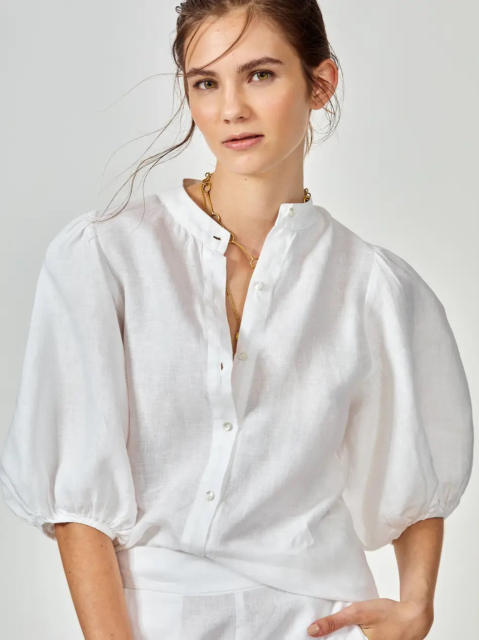 A woman wearing a white button-up blouse with puffed three-quarter sleeves and gold necklaces stands against a plain light background, looking slightly to the side.