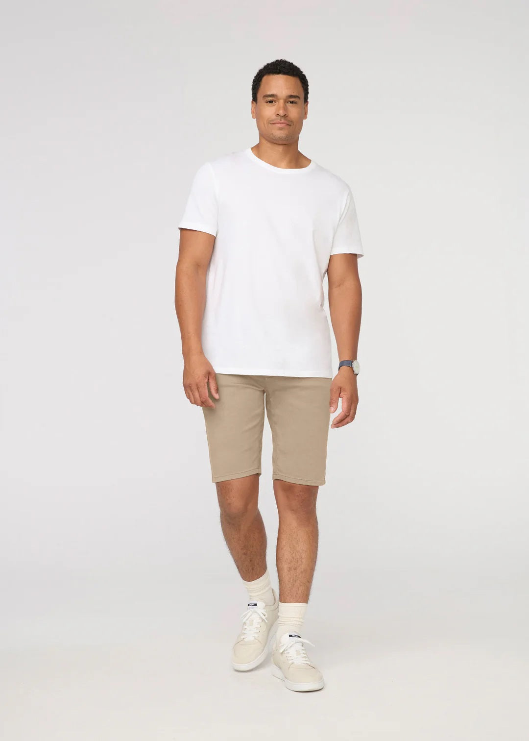 A person wearing a white t-shirt, DUER No Sweat Relaxed Shorts in Desert Khaki, white crew socks, white sneakers, and a wristwatch stands against a plain light background.