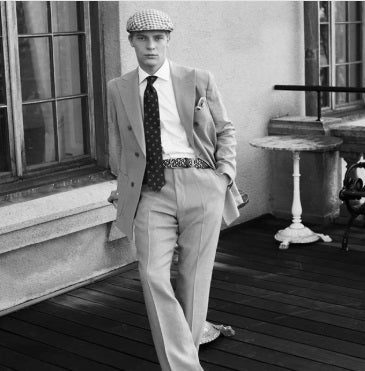 A man in a double-breasted suit, patterned tie, and WIGENS SIGNATURE WOVEN BELT IN GREY, BROWNS AND BLACK by WIGENS with a flat cap leans against a windowsill on a wooden patio in this vintage black-and-white photo.