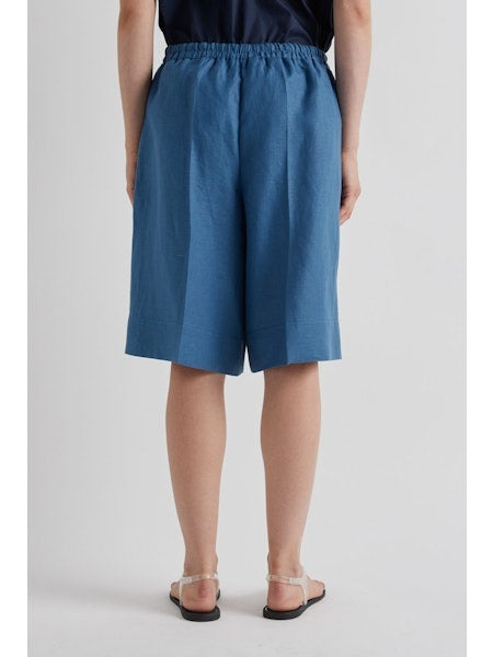 A person wears MEIMEIJ's Pull On Linen Short in Cerulean featuring a loose fit, knee length, and elastic waist, styled with flat strappy sandals against a plain light-colored background.