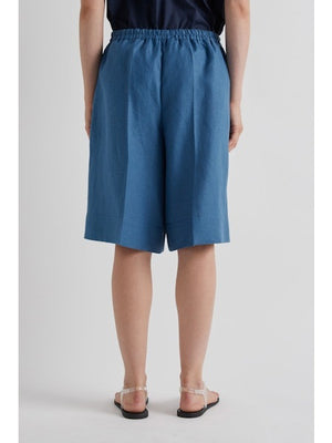 A person wears MEIMEIJ's Pull On Linen Short in Cerulean featuring a loose fit, knee length, and elastic waist, styled with flat strappy sandals against a plain light-colored background.