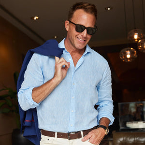 A man with short brown hair sits indoors, wearing the COLLARS AND CO - TOSCANO LINEN SHIRT IN LIGHT BLUE and white pants, resting his arm on a chair’s armrest and looking at the camera with a relaxed expression.