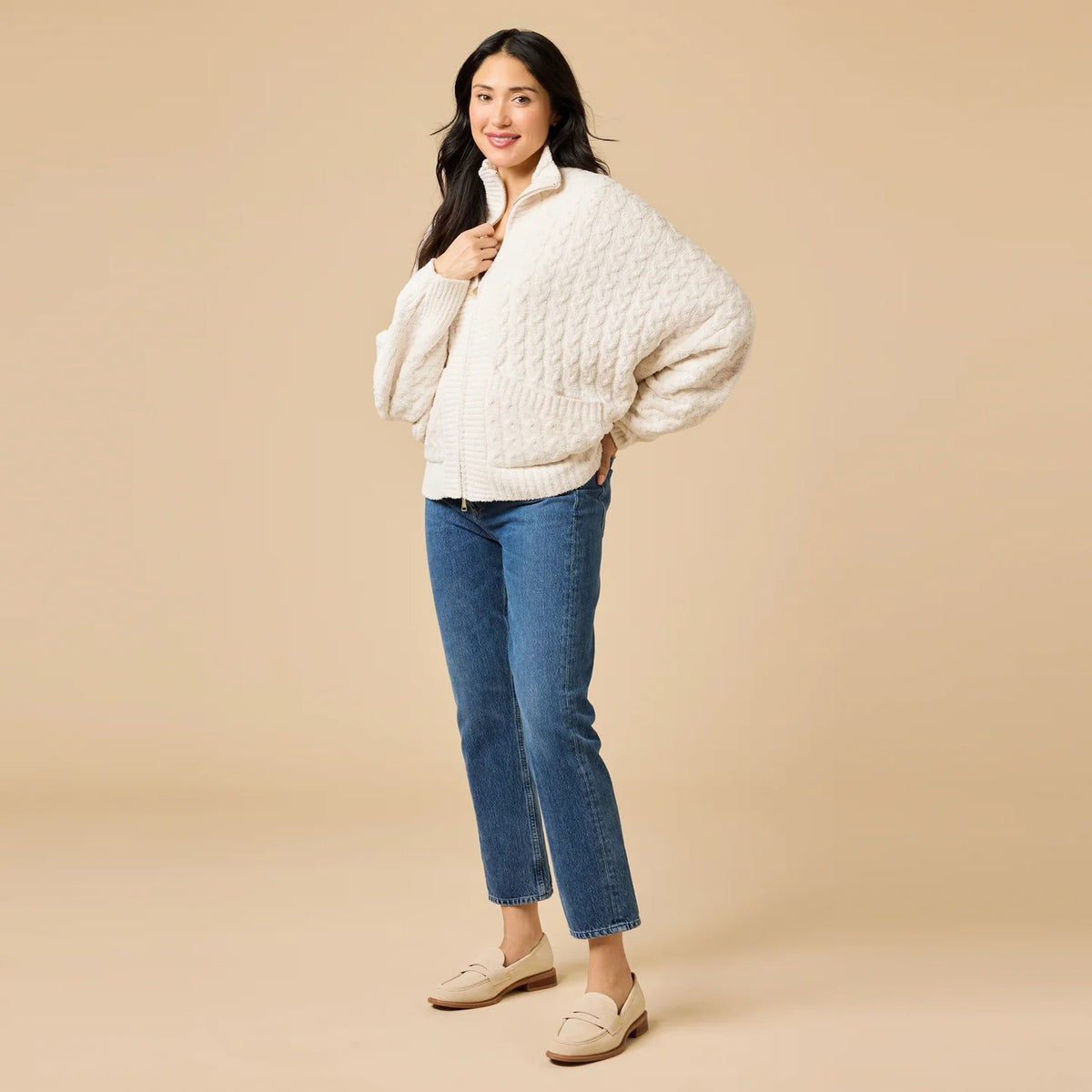 A woman wearing a cream-colored, plush SOFTIES - CABLE KNIT MARSHMALLOW FUNNEL NECK DOUBLE ZIP JACKET with a funnel neck and double zipper, paired with blue jeans.