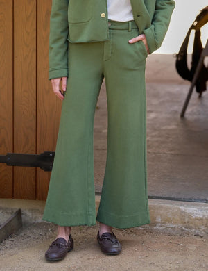 A woman stands by a wooden stable door, wearing the FRANK & EILEEN Riley Trouser Sweatpant in Nori from the California Heritage Collection, styled with a matching green jacket, white shirt, and dark loafers. She looks down as she gently touches her hair, while a saddle and tack hang in the background.