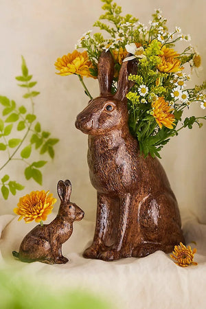 The QUAIL CERAMICS - HARE VASE, LARGE by QUAIL DESIGNS is a hand-painted brown ceramic vase shaped like a hare, displayed with yellow and white flowers and greenery. A single yellow flower lies beside it on a white background.