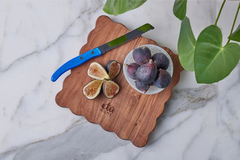The ETUHOME Natural Square Scalloped Cutting Board, Medium, crafted from durable acacia wood, stands upright against a light gray wall on a white marble surface.
