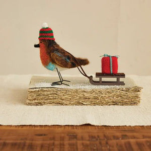 The CREATIVE COOP FELT ROBIN PULLING SLEIGH features a handmade wool felt robin in a red and green knit hat beside a sled with a red gift—perfect as festive holiday decor or a unique gift.