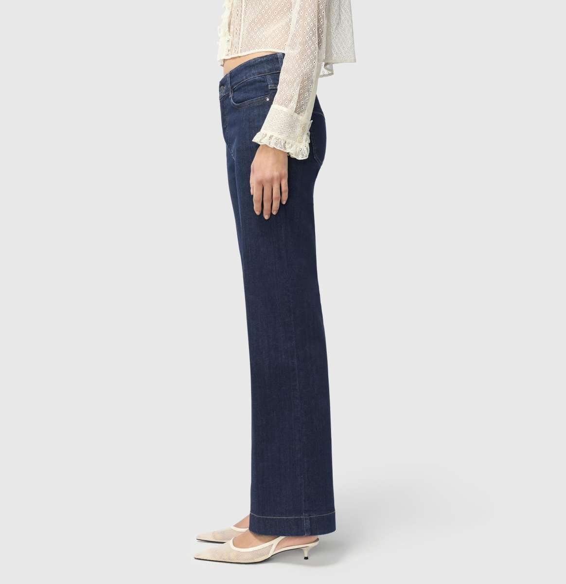 A person with light brown hair stands against a plain background, wearing a sheer, ruffled, long-sleeved white blouse and MAC JEANS - DREAM WIDE AUTHENTIC by MAC in dark blue. They&#39;re paired with comfortable white shoes.
