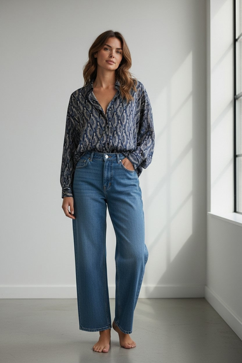 A woman with wavy hair stands barefoot indoors, looking at the camera and posing with one hand in her pocket. She wears high-waisted wide-leg blue jeans paired with the HARTFORD COSMA Long Sleeve Shirt in Blue Peacock Print by HARTFORD, featuring a boxy cut and subtle patterned design.